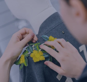 The image shows a person's hands embroidering a vibrant yellow flower design on a grey textile garment or fabric