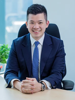 Bing Xi Sia, a smiling young Asian man in a navy blue suit and patterned tie, sits at a desk in a professional office setting