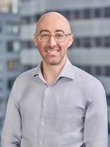 A smiling middle-aged man with glasses and a beard, wearing a light gray button-up shirt, stands in front of a blurred office building backdrop
