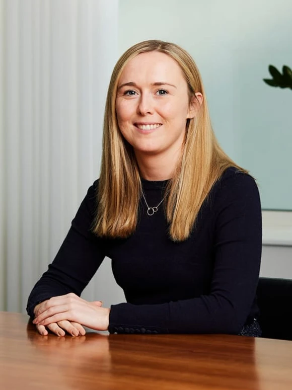The image shows a smiling woman with long blond hair, dressed in a black top, sitting at a desk and looking directly at the camera