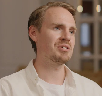 A close-up portrait of a man with blond, wavy hair and a short beard, wearing a white shirt and looking intently at the camera