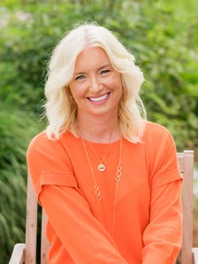 The image shows Carolyn Everson, a smiling woman with blonde hair, wearing an orange sweater and necklaces, surrounded by lush greenery