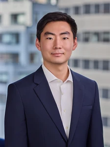 The image shows Daniel Lee, a young Asian man in a navy blue suit and white shirt, standing in front of a modern building and looking directly at the camera with a neutral expression