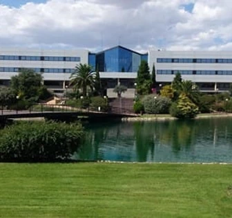 A modern university campus with a large multi-story building, landscaped gardens, and a tranquil reflecting pool in the foreground
