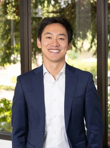 A smiling Asian man in a navy blue suit standing in front of a window, looking happy and confident. This is Andrew Ren