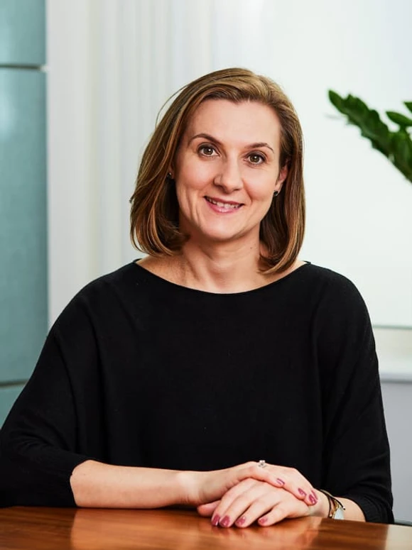 A smiling woman with brown hair, wearing a black top, sitting at a desk in a professional setting