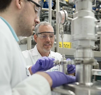 Two lab technicians in white coats and safety glasses work on equipment in a pharmaceutical manufacturing facility