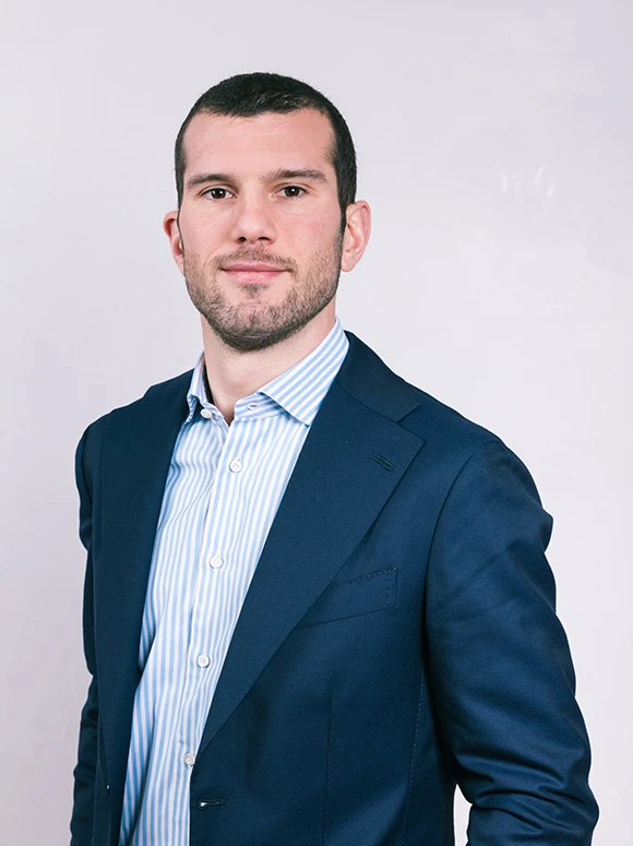 The image shows a young man with dark hair, wearing a navy blue suit and a striped dress shirt, looking directly at the camera with a serious expression