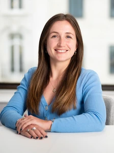 The image shows Anna Mason, a smiling woman with long brown hair, wearing a blue sweater and sitting at a desk in a professional setting