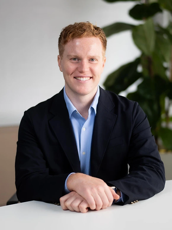 A smiling man with reddish-brown curly hair, wearing a dark blue suit and collared shirt, confidently sits at a table