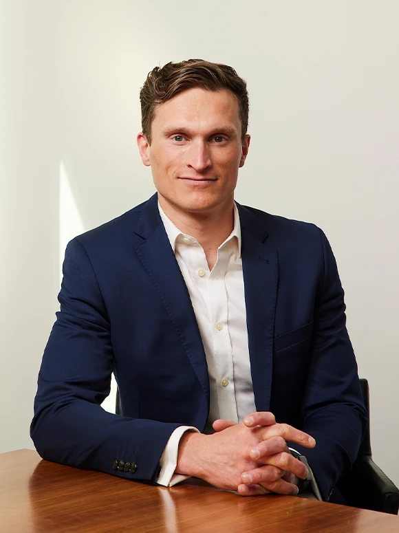 The image shows a confident middle-aged man wearing a navy blue suit sitting at a desk, hands folded, with a warm and friendly expression on his face