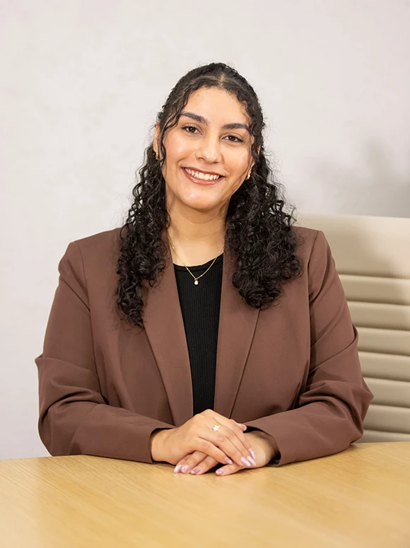 Adriana Ochoa, a smiling woman with long, curly dark hair, wearing a brown jacket, seated at a table and looking directly at the camera
