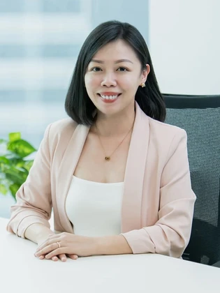 The image shows Evangeline Sim, a smiling young professional woman in a beige blazer, seated at a desk with a blurred background