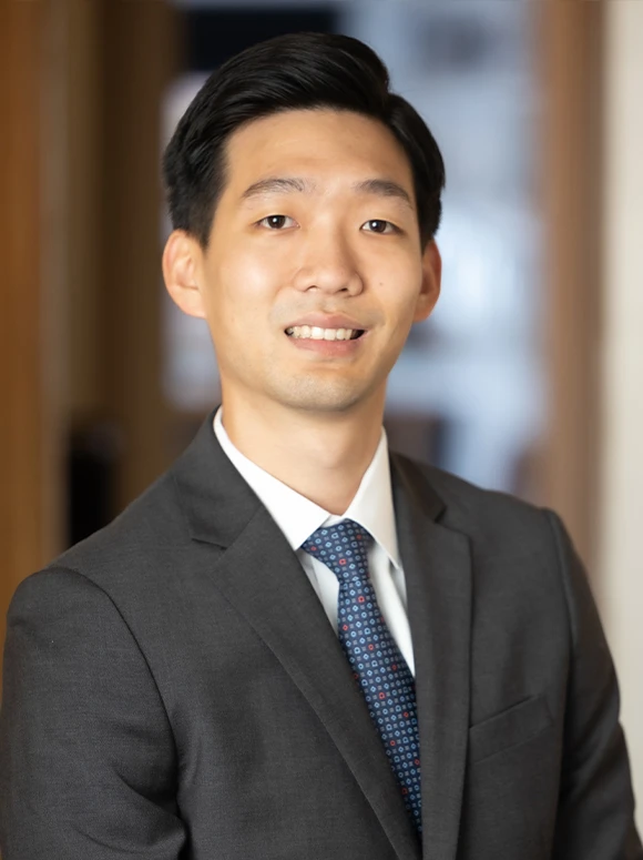 The image shows a smiling Asian man in a dark suit and tie, posing for a professional portrait