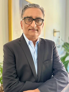 Raj Patel, a man in a business suit with glasses, sitting in an indoor setting with a hopeful expression