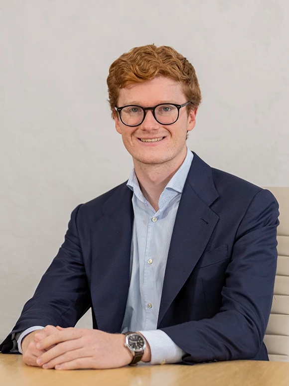 The image shows a smiling young man with curly red hair wearing glasses and a navy blue suit, sitting at a desk. The filename indicates his name is Jaime Tejedor