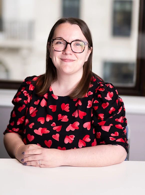 The image shows a smiling woman wearing glasses and a red heart-patterned blouse, sitting at a table and looking directly at the camera