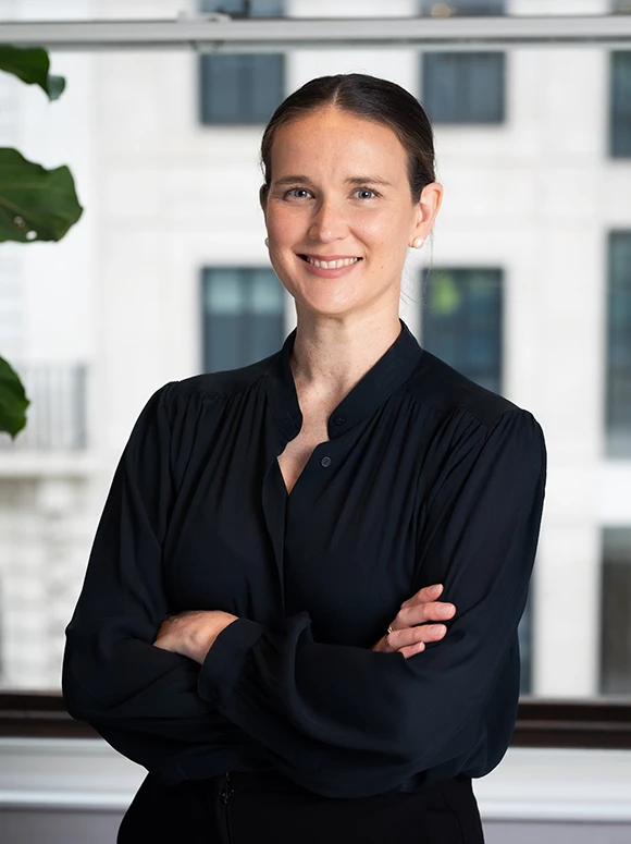 The image shows Jessica Haigh, a woman in a professional black blouse, standing in front of a background of office windows and smiling at the camera