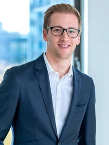 Fabian Selzam, a smiling, bespectacled man in a navy suit, standing in front of a large window with a professional, business-like demeanor