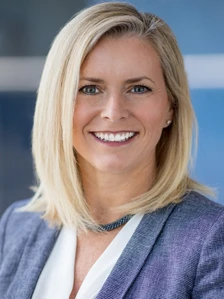 The image shows a smiling, middle-aged woman with long, blonde hair wearing a gray blazer against a blue background. The filename suggests this is a headshot photo of Kelly