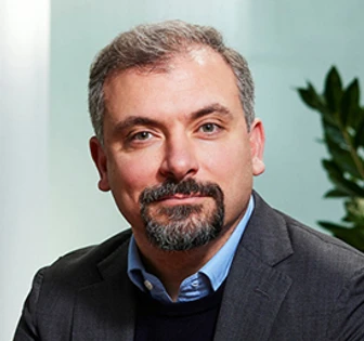 The image shows a middle-aged man with gray hair and a beard, dressed in a suit and looking directly at the camera with a serious expression