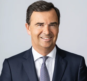 The image shows a smiling middle-aged man in a navy suit and patterned tie, with short dark hair, against a plain gray background. The filename indicates his name is Christian Ulbrich