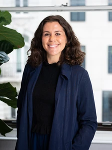 The image shows a smiling woman with curly brown hair wearing a navy blue coat, standing in an office environment with plants in the background