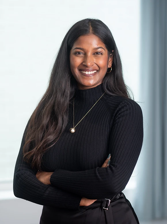 The image shows a smiling woman with long dark hair, wearing a black turtleneck sweater and a pearl necklace. She appears to be in her early to mid 40s and has an open, friendly expression