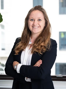 The image shows Apolline Tirot, a smiling woman with long, curly brown hair, dressed in a professional black suit, posing confidently in a modern office setting