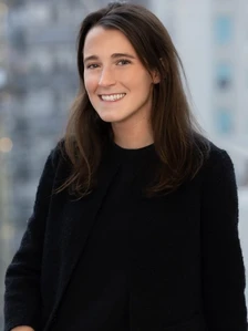 The image shows a smiling young woman with long brown hair wearing a black sweater, standing outdoors in front of a blurred background
