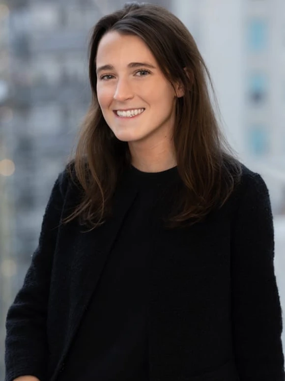The image shows a smiling young woman with long brown hair wearing a black sweater, standing outdoors in front of a blurred background