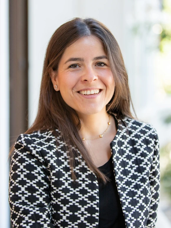 The image shows a smiling woman with long dark hair wearing a black and white patterned blazer, posing for a professional headshot