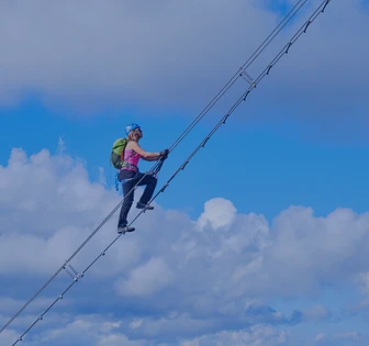 A person wearing a harness and helmet is suspended on a cable high in the sky against a backdrop of blue sky and clouds, engaging in an aerial adventure activity