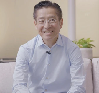 The image shows a smiling Asian man in a white dress shirt, wearing round glasses, sitting in a room with a potted plant visible in the background