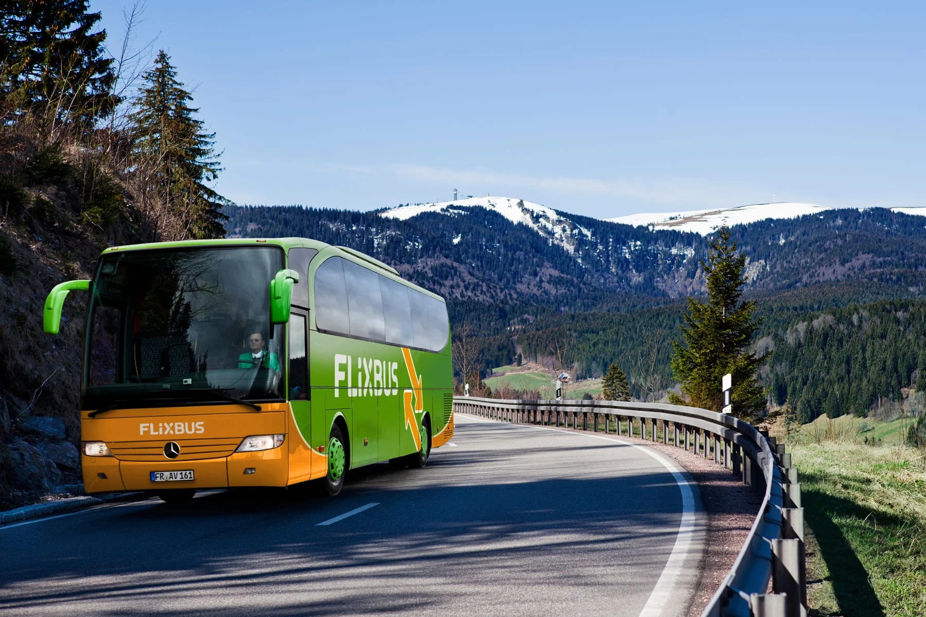A green and orange Flixbus coach traveling on a scenic mountain road surrounded by snow-capped peaks and pine trees