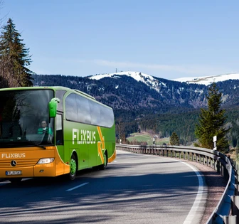 A green and orange Flixbus coach traveling on a scenic mountain road surrounded by snow-capped peaks and pine trees
