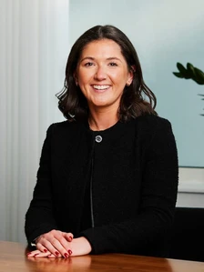 The image shows Harriet Neale, a woman with dark hair and a warm smile, sitting at a desk and wearing a black jacket