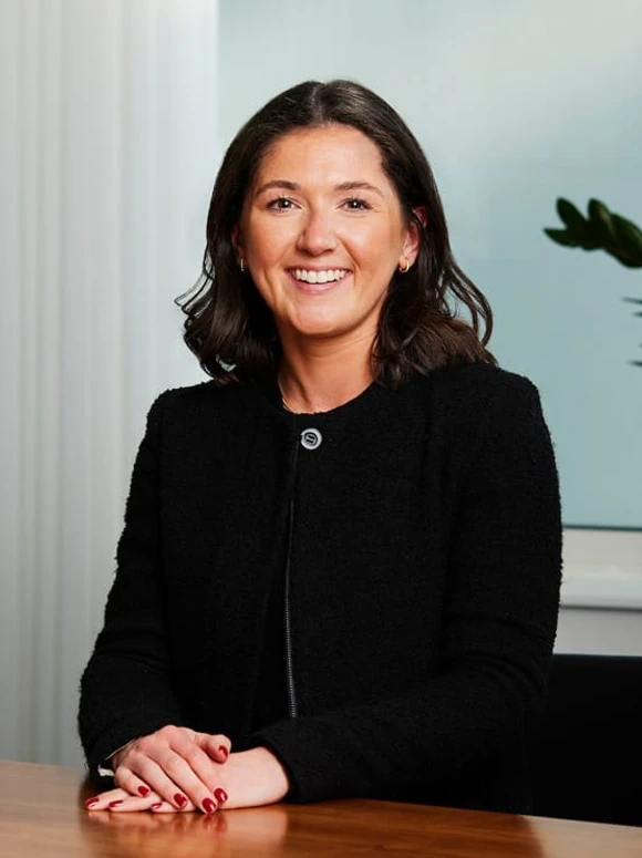 The image shows Harriet Neale, a woman with dark hair and a warm smile, sitting at a desk and wearing a black jacket