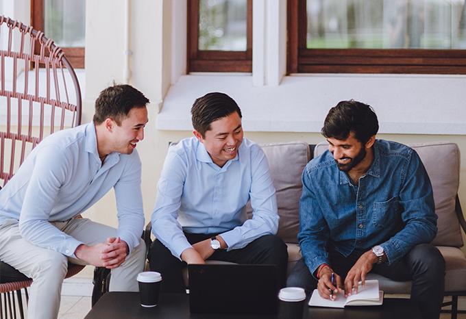 Three men in business casual attire are seated on a couch, engaged in conversation and taking notes in a bright, modern office space
