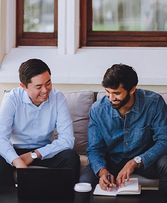 Three men in business casual attire are seated on a couch, engaged in conversation and taking notes in a bright, modern office space