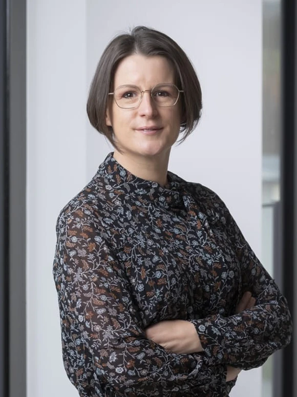 The image depicts Julie Schleich, a woman in her 40s or 50s, wearing a floral patterned top and eyeglasses, standing with her arms crossed and a serious expression on her face