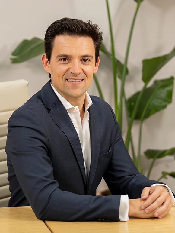 The image shows a smiling man wearing a navy blue suit, seated at a desk with potted plants in the background. The man appears to be Alvaro Marin, a professional individual