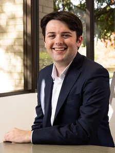 A man with brown hair and a warm smile, wearing a navy suit, sitting at a desk in an office setting. The filename indicates the man's name is Conor Holahan