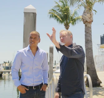 Two men, one in a white shirt and the other in a navy jacket, standing near a marina with palm trees in the background