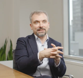 The image shows Riccardo Basile, a middle-aged man with gray hair and a beard, sitting at a desk and gesturing while wearing a suit