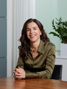 Silvia Oteri, a middle-aged woman with dark curly hair, is smiling warmly while sitting at a desk in a professional setting