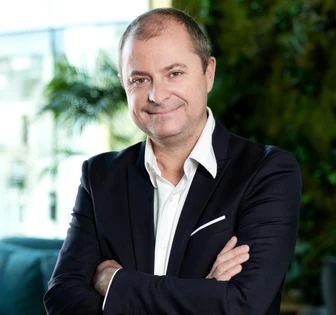 Antoine, a smiling middle-aged man in a black suit, stands confidently with his arms crossed in a professional office setting