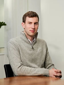 A man in a gray sweater, with short brown hair, sitting at a desk and looking directly at the camera