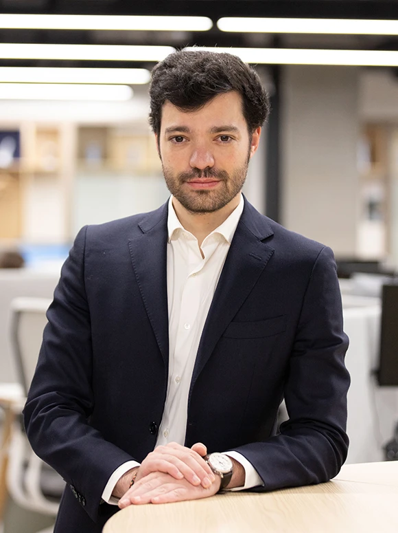 The image shows Javier Vasquez, a professional-looking man in a navy blue suit, sitting at a desk with a focused expression