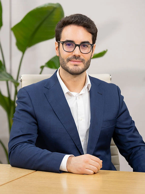 The image shows Julio Palomares, a professional-looking man with dark hair and glasses, wearing a blue suit, sitting at a desk with greenery in the background